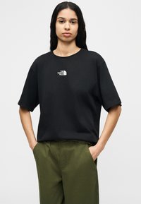 Γυναίκα με μακριά σκούρα μαλλιά φοράει μαύρο μπλουζάκι The North Face και πράσινο παντελόνι, στέκεται με τα χέρια στις τσέπες μπροστά σε μονόχρωμο φόντο.