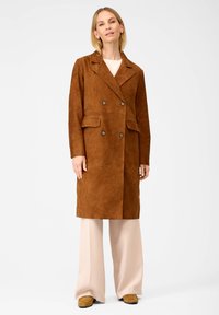 Brauner doppelseitiger Wildledermantel mit aufgesetzten Taschen, Reverskragen und Knopfakzenten, kombiniert mit hellfarbigen, weit geschnittenen Hosen.