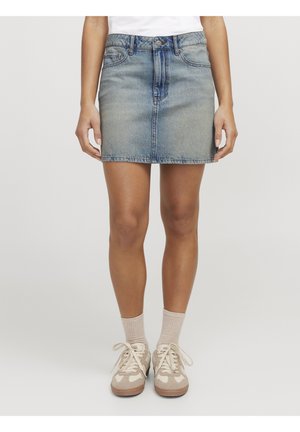 Person iført en blå denim mininederdel, beige sokker og beige og hvide snøresneakers, stående på en ensartet lysegrå baggrund.