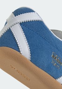 Sneaker in camoscio blu con accenti in pelle bianca, cuciture a zigzag e suola in gomma marrone testurizzata. Presenta il logo dorato "adidas TOKYO".