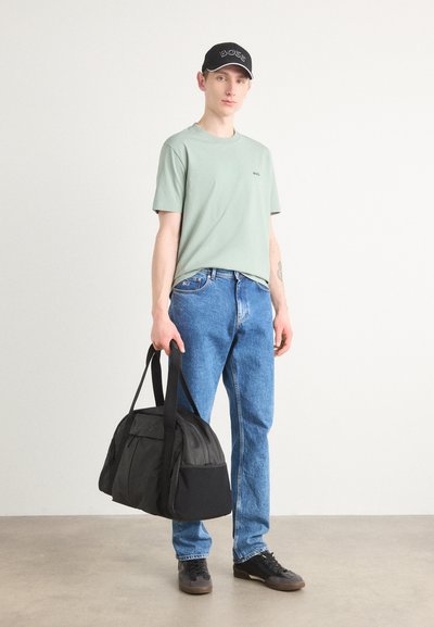 T-shirt vert clair à manches courtes, jeans en denim bleu, sac noir avec logo, et chaussures noires. Le modèle se tient sur un fond neutre.