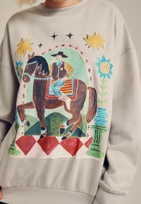 Szürke pulóver, amelyen egy színes illusztráció látható egy férfiról, aki lovagol, virágmintákkal és gyémánt mintázatú kerettel díszítve.