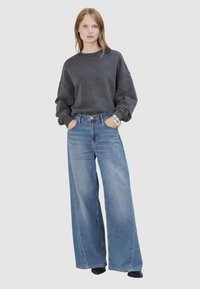 Frau trägt übergroßen, ausgewaschenen schwarzen Sweatshirt, weit geschnittene blaue Jeans und silbernes Armband, steht mit den Händen in den Taschen vor einem grauen Hintergrund.