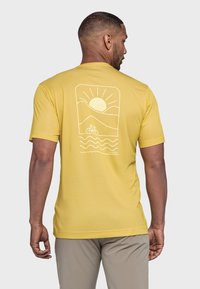 Gelbes Baumwoll-T-Shirt mit einem weißen Grafikdesign auf der Rückseite, das einen Sonnenuntergang, Berge und ein Fahrrad über wellenförmige Linien darstellt.