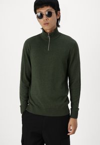 Pull à col zippé vert foncé en tissu doux, avec des poignets et un ourlet côtelés. Porté avec un pantalon noir et des lunettes de soleil.