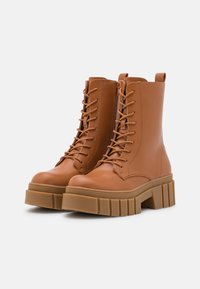 Botas de tornozelo em couro castanho com frente de atacadores, bico redondo e sola plataforma robusta. Apresenta um fecho de correr lateral e uma alça na parte de trás.