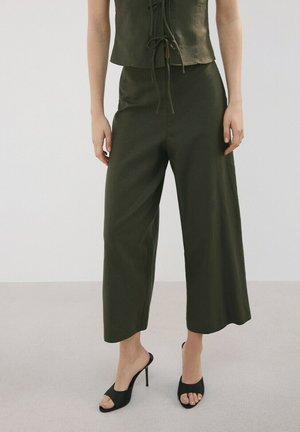 Femme portant un pantalon large court couleur olive et un haut sans manches assorti, associé à des mules noires à talons ouverts.