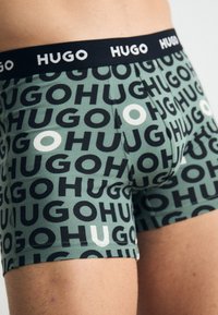 Grüne Boxershorts mit einem wiederholten Logo-Design in Schwarz und Weiß, ausgestattet mit einem schwarzen elastischen Bund, der mit "HUGO" beschriftet ist. Aus weichem Baumwollmaterial.