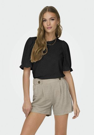 Ung kvinde med langt lyst brunt hår iført en sort top med pufærmer og højttaljede beige shorts med knapper.