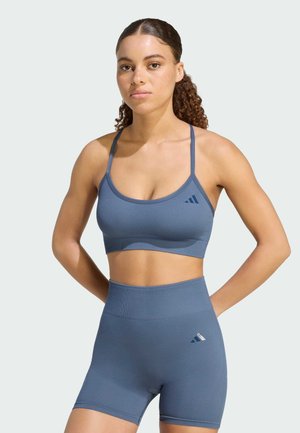Mujer de pie con las manos detrás de la espalda, vestida con un sujetador deportivo azul a juego y pantalones cortos de ciclista de cintura alta con logotipos en el lado izquierdo.
