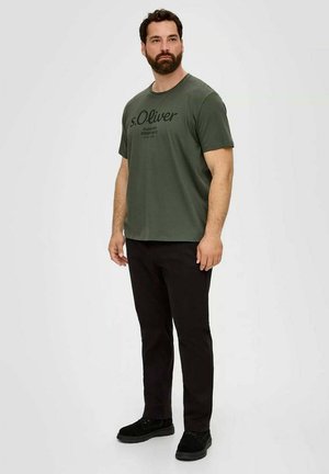 Man with beard wearing olive green s.Oliver graphic t-shirt, black pants, and black shoes standing against plain light background.