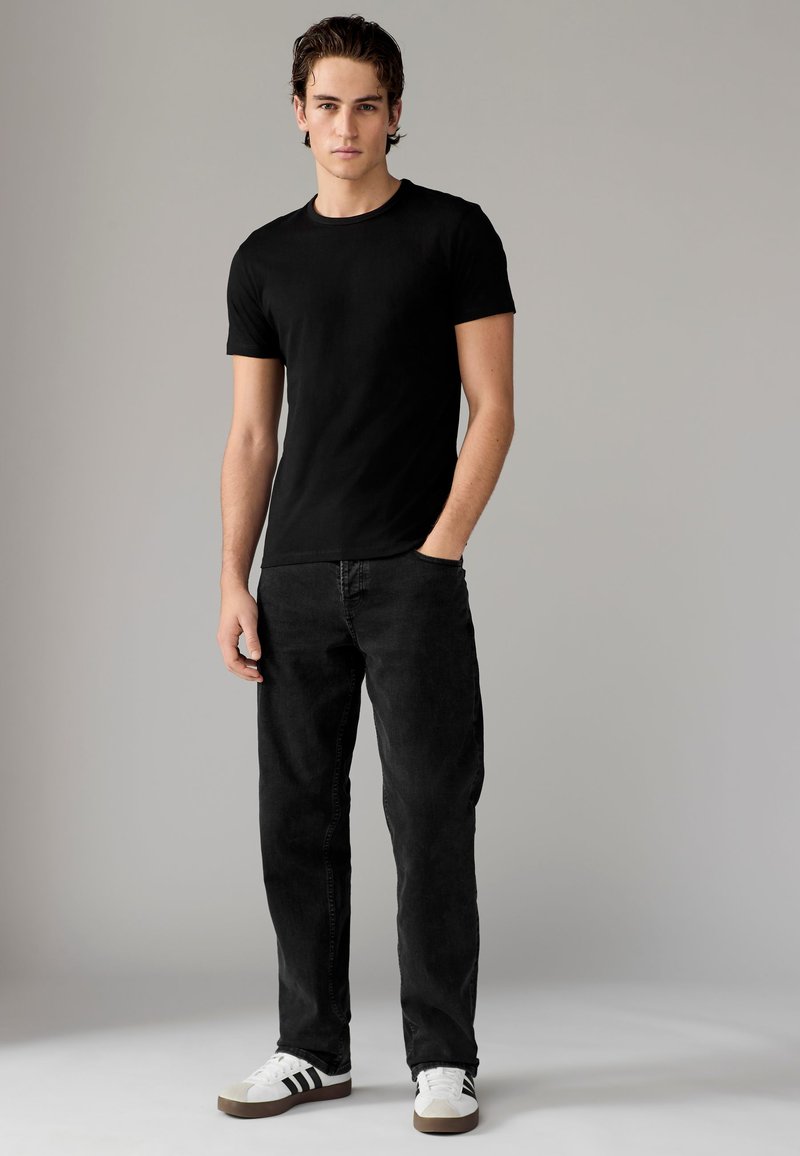 T-shirt nera in cotone a maniche corte, abbinata a jeans neri e scarpe da ginnastica bianche con strisce nere, in piedi su uno sfondo neutro.