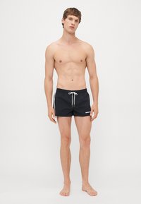 Ung man som står barfota och har på sig svarta badshorts med vit dragsko och sidostråk på en enkel bakgrund.