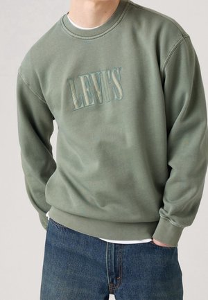 Osoba ubrana w zieloną bluzę Levi's z wyszywanym logo i niebieskie dżinsy, z rękami częściowo w kieszeniach, stojąca na jednolitym tle.