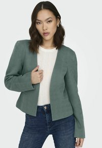 Teal visgraat blazer met een open voorkantontwerp, lange mouwen en subtiele textuurstof, gecombineerd met een licht topje en donkere jeans.
