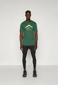 Das grüne Nike Trail T-Shirt mit kurzen Ärmeln verfügt über ein gelbes Logo und ein Bergdesign; kombiniert mit schwarzen Leggings und Sportschuhen.