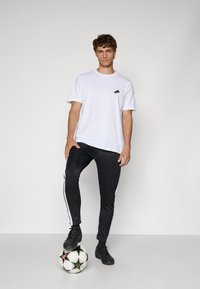 Hvid t-shirt med korte ærmer og et sort logo på brystet, parret med sorte sweatpants med hvide sidestreaks. Sorte fodboldstøvler.