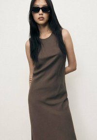 Abito marrone senza maniche con un design morbido e aderente. Modella che indossa occhiali da sole neri eleganti. Sfondo semplice, che mette in risalto l'outfit.