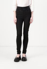 Černé skinny džíny s vysokým pasem, zapínání na dva knoflíky, přední kapsy a hladký textilní povrch. Nosené s černými platformovými botami.
