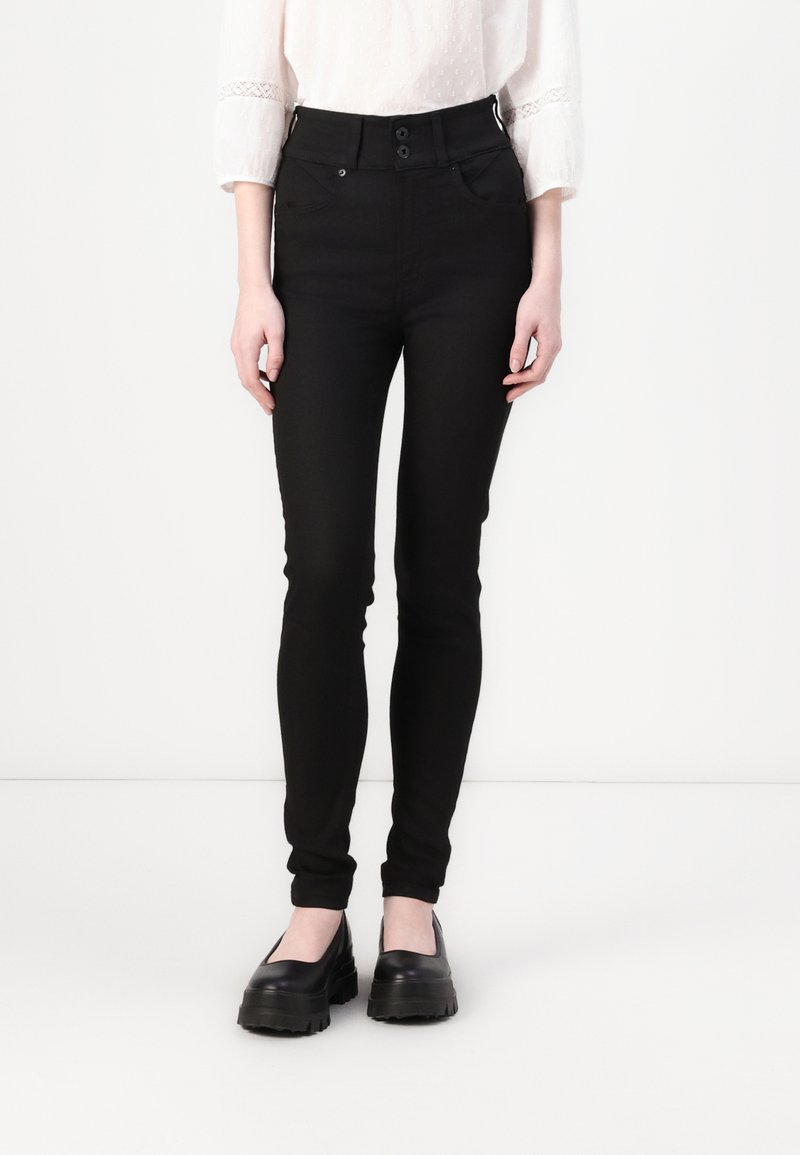 Černé skinny džíny s vysokým pasem, zapínání na dva knoflíky, přední kapsy a hladký textilní povrch. Nosené s černými platformovými botami.