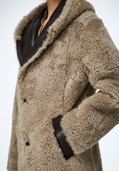 Palton bej din blană falso cu accente din piele maro, prevăzut cu un glugă mare, design cu două rânduri de nasturi și buzunare laterale. Suprafață moale, texturată.