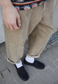 Una persona che indossa pantaloni in velluto a coste e scarpe nere senza lacci si trova su un pavimento decorato, con calze bianche in vista.