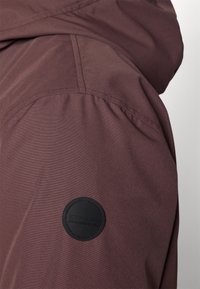 Veste en tissu marron avec une texture lisse, dotée d'un patch logo rond noir sur la manche et de détails de couture visibles.