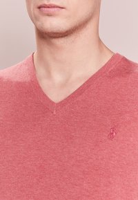 Homme portant un pull rose à col en V avec un petit logo brodé sur le côté gauche de la poitrine, visible du cou au haut du torse.