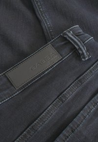 Tamsaus džinsinio audinio kelnės su lygiu paviršiumi ir odiniu ženkleliu su įspaustu užrašu "GABBA". Matomas siūlių dekoravimas.