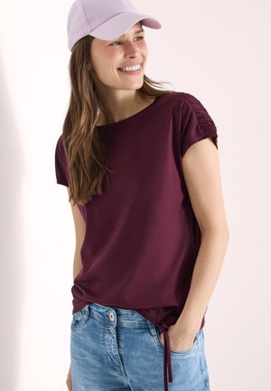 Femme souriante, portant une casquette violet clair, un haut à manches courtes bordeaux avec des manches froncées, et un jean en denim bleu clair, une main dans la poche.