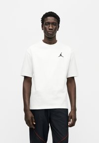 Λευκό βαμβακερό t-shirt με κοντά μανίκια, στρογγυλή λαιμόκοψη και μαύρο λογότυπο Jumpman στο αριστερό μέρος του στήθους. Φορεμένο με μπλε παντελόνια με κόκκινες λεπτομέρειες.