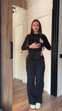 Γυναίκα με μακριά καστανά μαλλιά, που φοράει μαύρο τοπ και παντελόνι, κρατώντας διπλωμένο μαύρο ύφασμα με σχέδια, στέκεται σε ξύλινο πάτωμα σε μοντέρνο δωμάτιο.