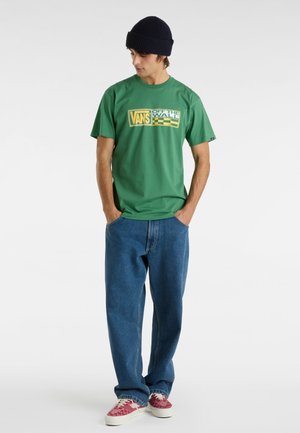 Camiseta verde de manga corta con un gráfico del logo de Vans, combinada con unos vaqueros azules holgados y zapatos rojos de estampado. Lleva puesto un gorro de punto azul marino.