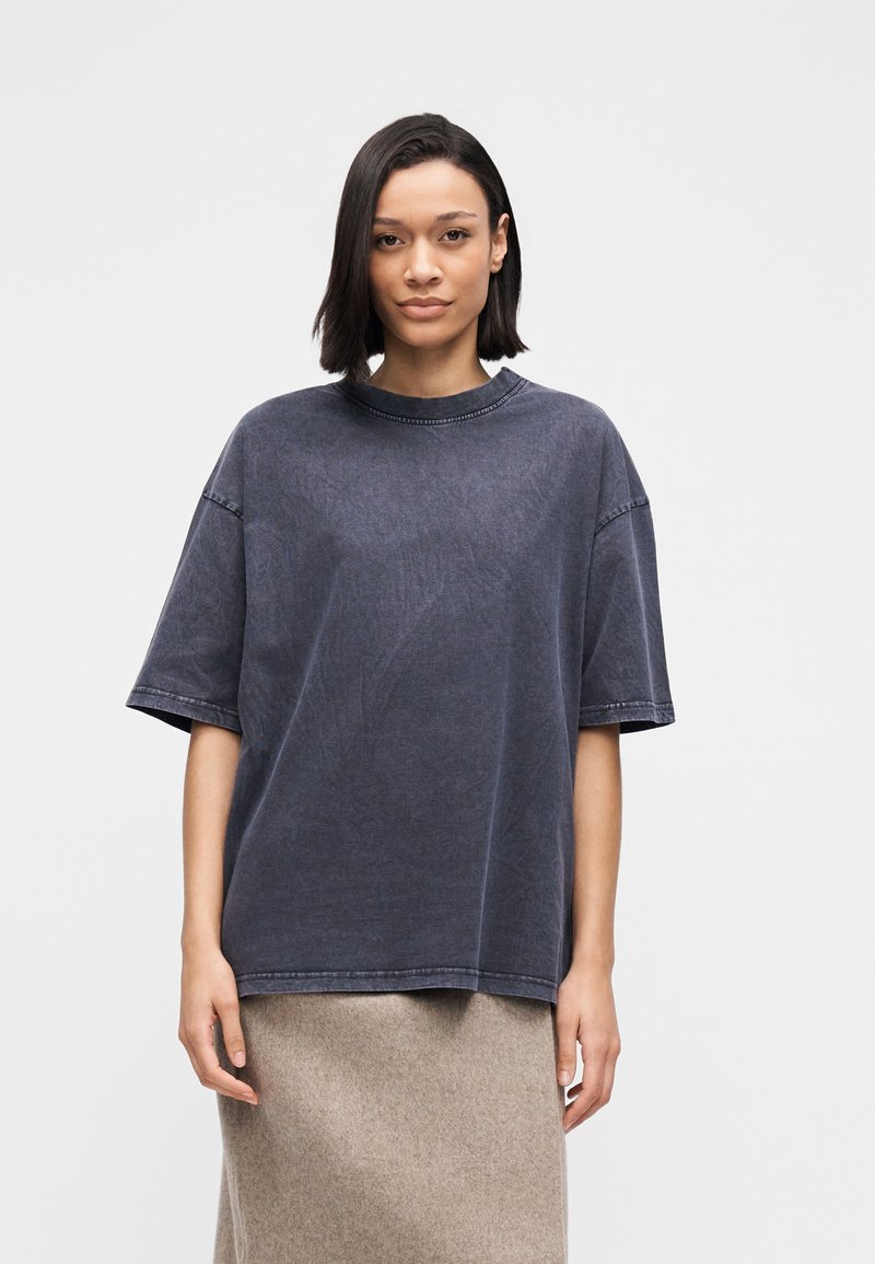 Γυναίκα με ίσια σκούρα μαλλιά που φοράει υπερμεγέθη σκούρο γκρι κοντομάνικο πουκάμισο και μπεζ φούστα, στέκεται μπροστά σε απλό ανοιχτό φόντο.