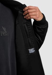 Giacca bomber nera con polsini a costine, dotata di uno strato interno in morbido tessuto e di un'etichetta cucita che mostra "Khujo™ original changing threads."