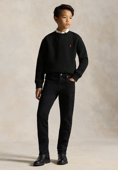 Băiat purtând un pulover negru, cămașă albă cu guler, blugi negri și cizme negre, stă cu o mână în buzunar pe un fundal neutru.