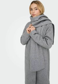 Cappotto grigio in lana con una grande sciarpa drappeggiata. Il cappotto presenta una vestibilità rilassata e cuciture minime, sottolineando un tessuto liscio e tattile.