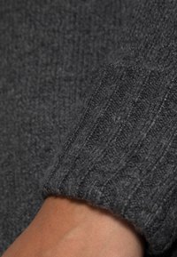 Cuff di un maglione grigio a coste, con una consistenza morbida e sottili variazioni di tonalità. Il bordo presenta una finitura leggermente arrotolata.