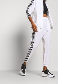 Ensemble de sport blanc avec accents à trois bandes noires, veste ajustée et courte, et pantalon à ceinture élastique. Baskets noires et blanches.