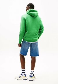 Grüne Kapuzenjacke, Denim-Shorts, weiße Sneaker mit blauen und gelben Akzenten sowie gestreiften Socken. Rückansicht, lässiger Stil.