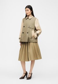 Γυναίκα που φοράει διπλόσαρκο μπεζ και καφέ σακάκι, πλισέ φούστα μέχρι το γόνατο και καφέ ψηλοτάκουνα παπούτσια Mary Jane, στέκεται μπροστά σε λευκό φόντο.