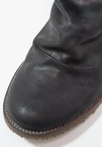 Bota de cuero negro con una textura ligeramente arrugada, punta redondeada y suela de goma marrón en contraste con costuras visibles.