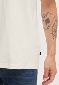 Hvid bomulds t-shirt med korte ærmer og en lige kant, kombineret med blå denimjeans. Synlig tatovering på armen viser spillekort.