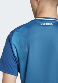Mėlyna sportinė marškinėlių su tekstūruotu gradientiniu dizainu, auksinėmis detalėmis ir žodžiu "SVERIGE" viršutinėje nugaros dalyje. Trumpomis rankovėmis ir apvaliu kaklu.