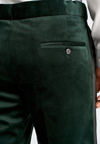 Close-up of dark green velvet trousers with a rear buttoned welt pocket and a person’s hand resting by their side.