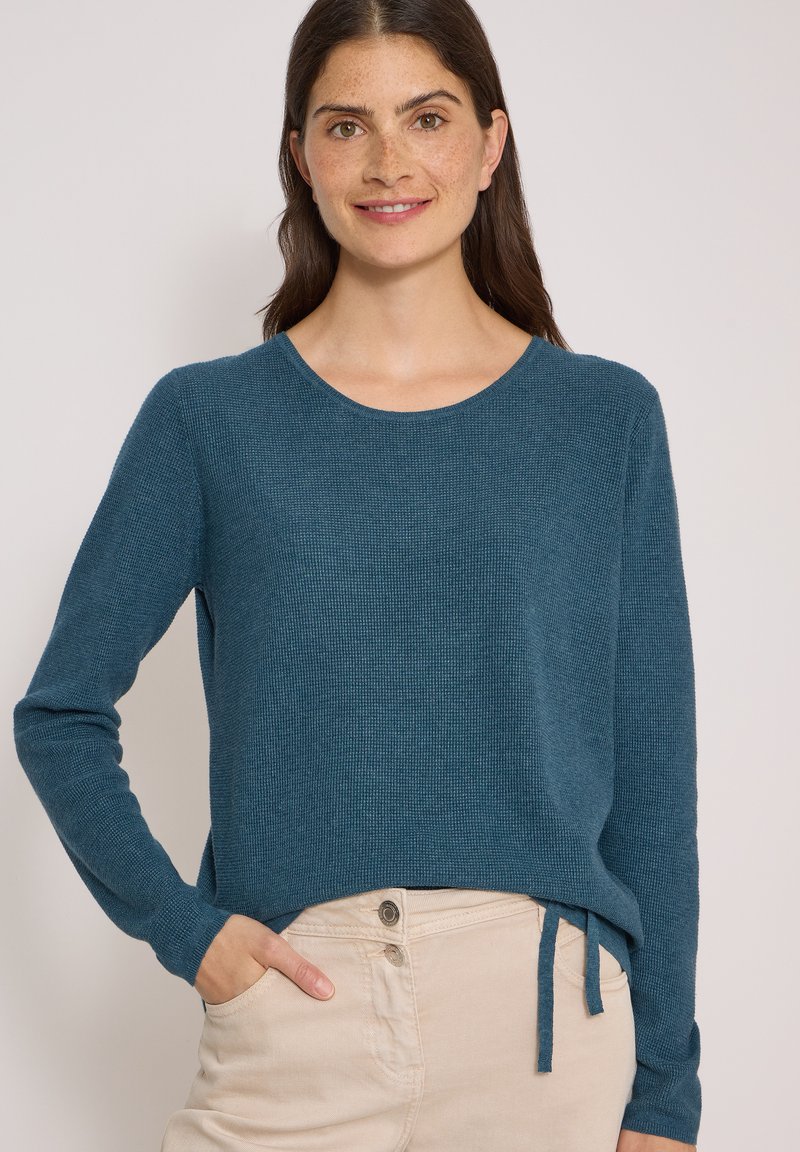 Femme souriante, portant un pull à manches longues couleur sarcelle avec un détail de lien et un pantalon beige clair, une main dans la poche.