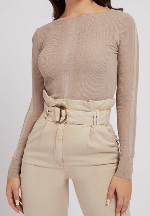 Kvinde iført en tætsiddende beige langærmet top og beige høj-taljede bukser med bælte og spænde detalje.