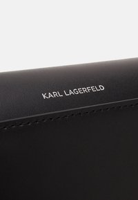 Musta nahkainen lompakko, jossa on tekstuuri pinta, sisältäen hopeanvärisen kohokuvioidun "KARL LAGERFELD"-logon ja ommeltuja yksityiskohtia reunoilla.