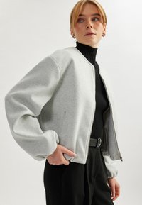 Harmaa pommikauppatakki, joka on valmistettu pehmeästä huovasta. Varustettu vetoketjulla, ribbaus hihansuissa ja rento istuvuus. Yhdistetty mustan korkeakauluspaidan kanssa.