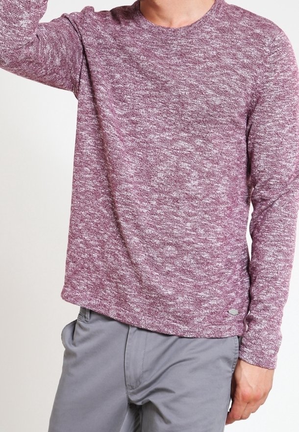 Personne portant un pull à manches longues bordeaux moucheté blanc avec un pantalon gris clair, bras légèrement levé hors cadre.